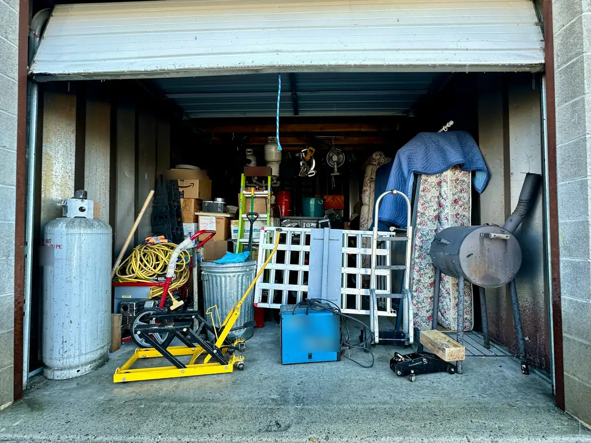 An open storage unit packed with tools, boxes, a hand truck, a smoker grill and a propane tank.